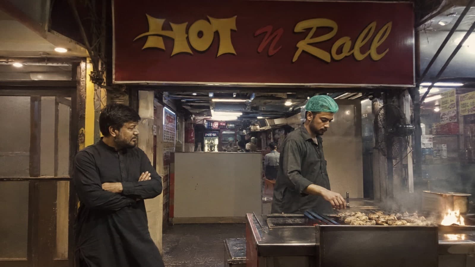 Hot n Roll's owner standing outside the storefront at night, arms crossed beside the glowing sign, while the chef grills skewers of tikka on the open flame behind him